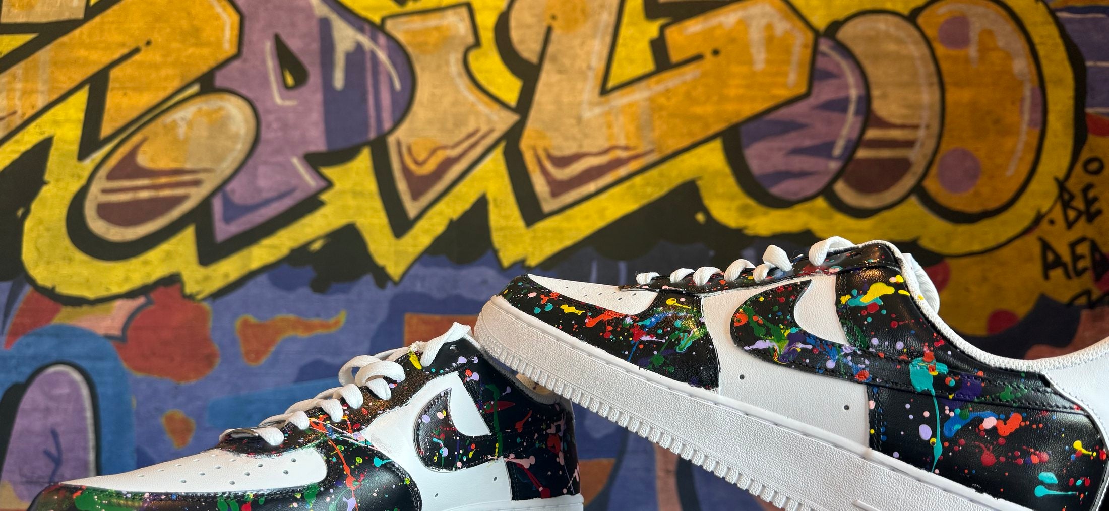 Both shoes of № 001 — hand-painted Nike Air Force 1 Lows, black panels covered in rainbow paint splatter, in front of a yellow graffiti wall