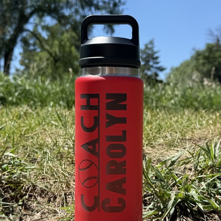 A red insulated water bottle engraved with COACH CAROLYN and a baseball, standing in grass