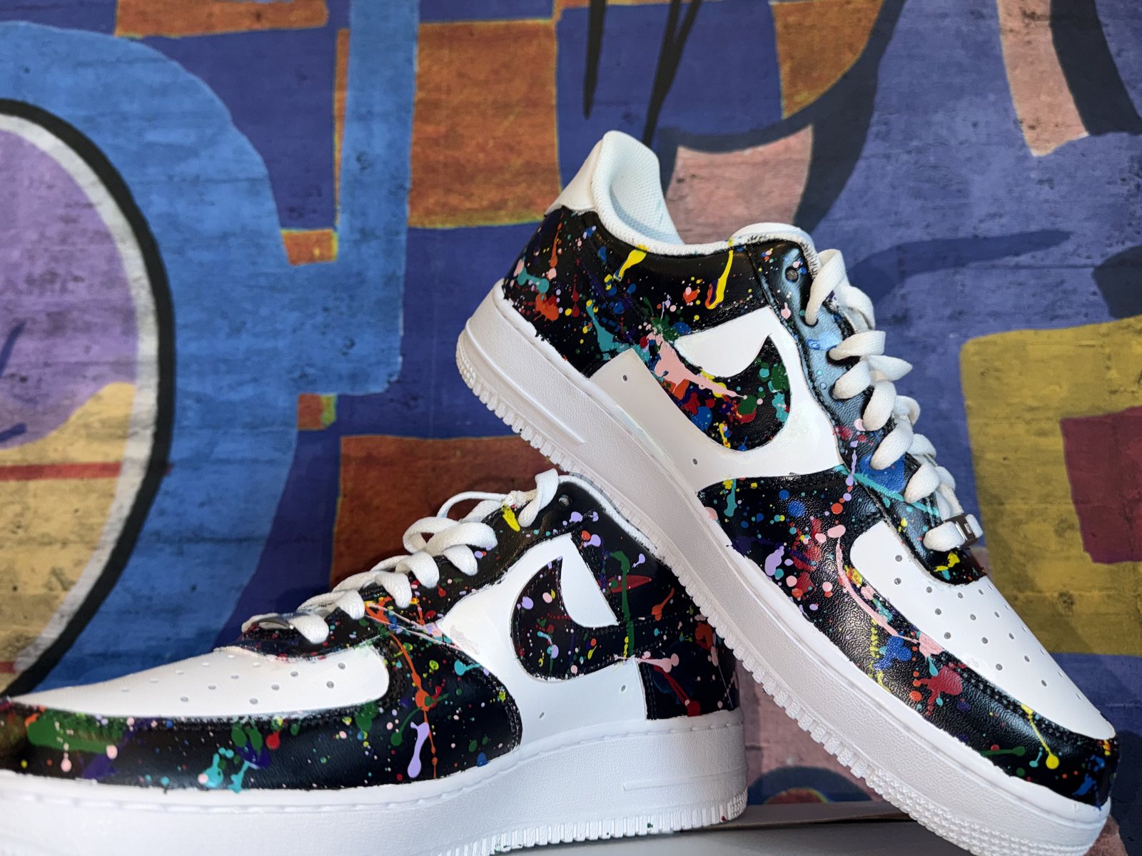 Hand-painted Nike Air Force 1s in black and white with rainbow paint splatter — № 001, full pair, against a blue graffiti mural